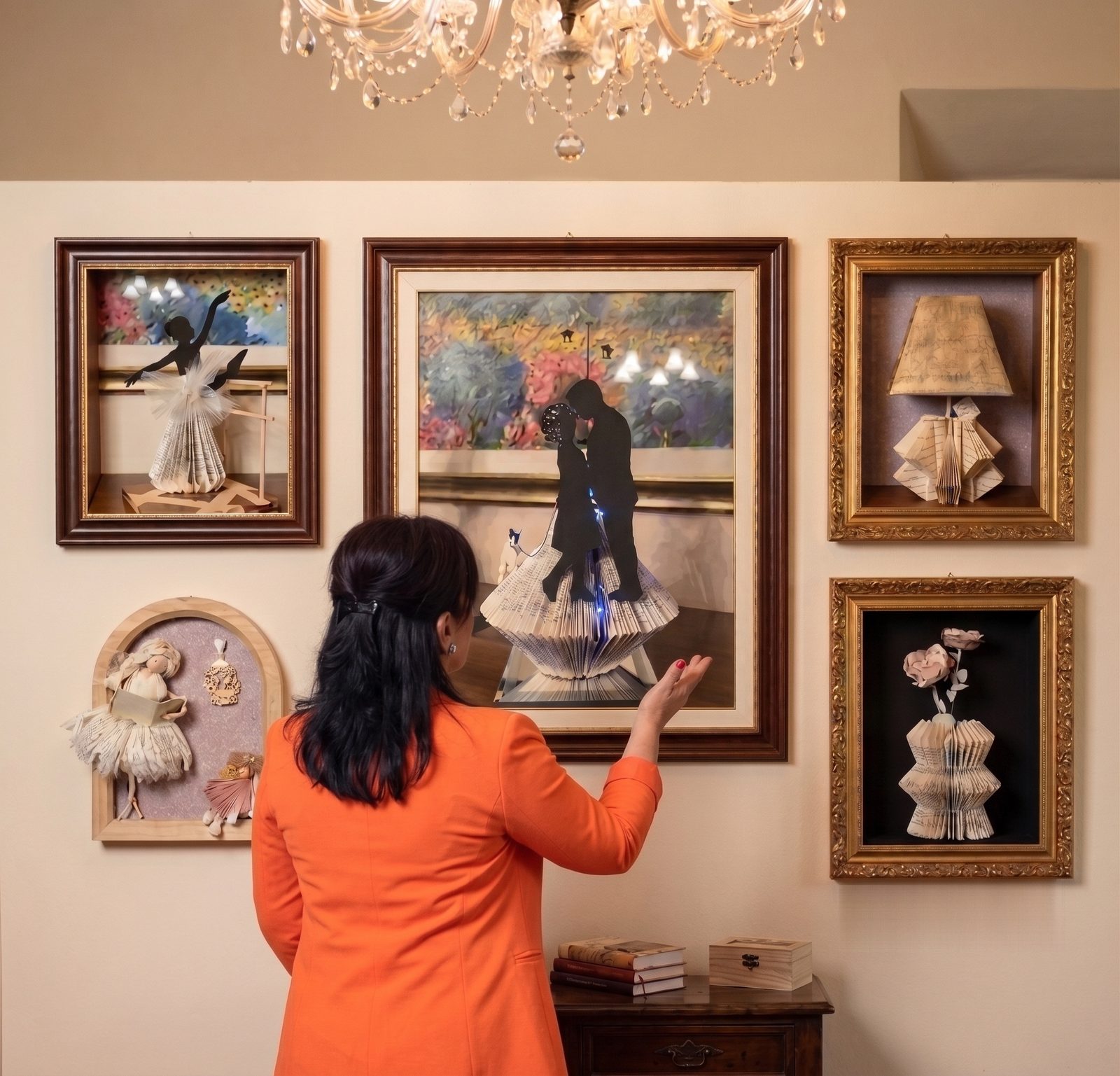 Antonella Peraio davanti alla parete delle sue opere, la mano tesa verso un quadro scultoreo: accanto lampade e bamboline in cornice, tutte ricavate da libri piegati a mano